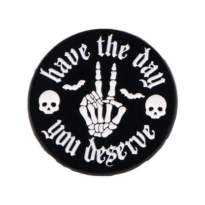 The Kiss Me Later round black enamel pin features a skeleton hand making a rude gesture, have the day you deserve in white text, plus spooky details like two skulls and two bats.