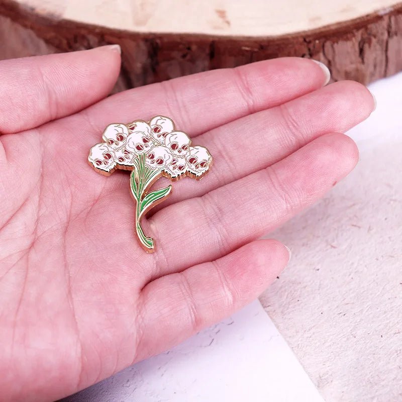 A person holds a brooch shaped like a Gothic Bouquet, featuring white flowers with green stems, gold outlines, and subtle hints of red roses in their palm.