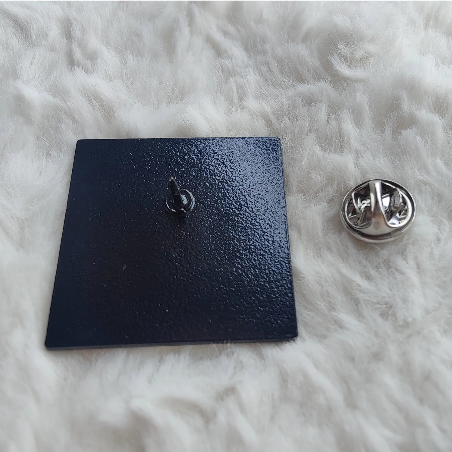 A black, square enamel badge pin by  featuring a pointed back and silver butterfly clutch, sits atop a soft white surface—a stylish accessory with Clueless-level charm.