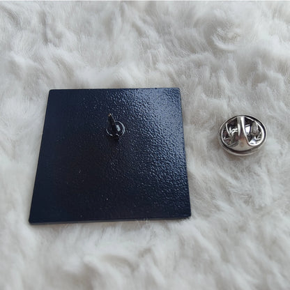 A black, square enamel badge pin by  featuring a pointed back and silver butterfly clutch, sits atop a soft white surface—a stylish accessory with Clueless-level charm.