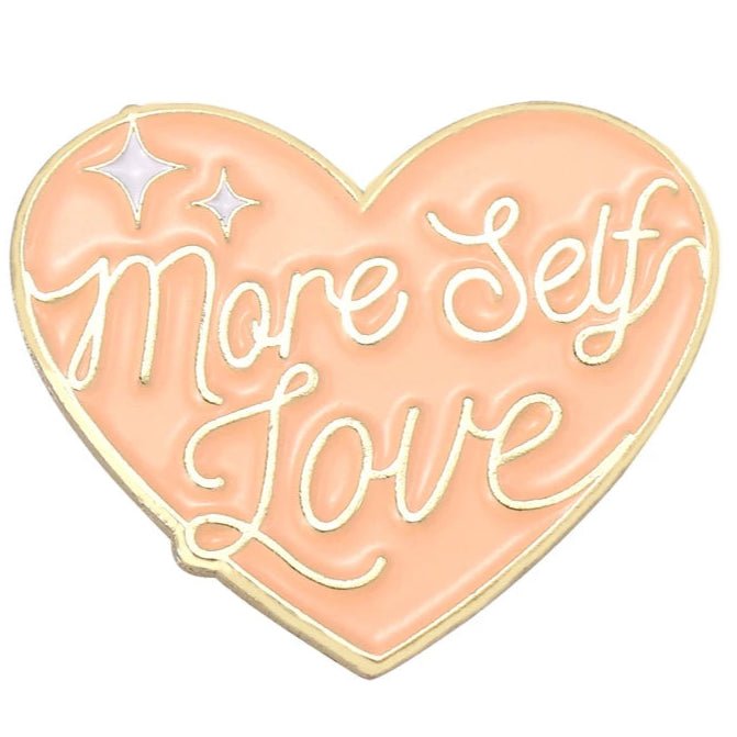 enamel pin is peach-colored in a heart shape with a gold outline, featuring More Self Love in handwritten text and two white twinkling stars on the left side.