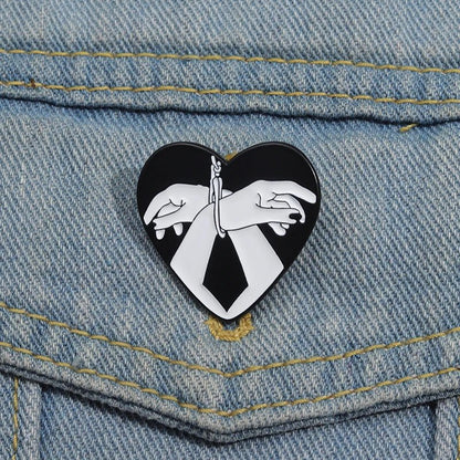 The Kiss Me Later Bound Hands enamel pin features two illustrated hands tied with string on a black love heart, making a bold statement accessory for your denim.