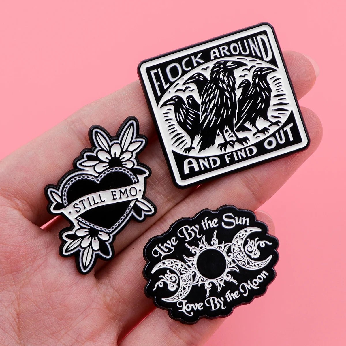 A hand holds three Kiss Me Later black-and-white enamel pins—Flock Around and Find Out (with crows), a heart reading Still Emo, and a sun & moon pin with Live By the Sun, Love By the Moon—adding spooky vibes against a pink background.