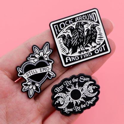 A hand holds three Kiss Me Later black-and-white enamel pins—Flock Around and Find Out (with crows), a heart reading Still Emo, and a sun & moon pin with Live By the Sun, Love By the Moon—adding spooky vibes against a pink background.