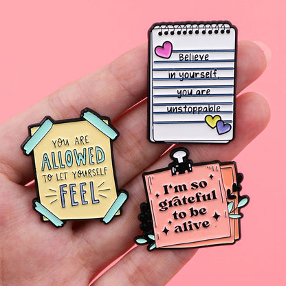A hand holds three colorful enamel pins with empowering affirmations—like You are allowed to let yourself feel and You are unstoppable—against a pink background, promoting mental health.