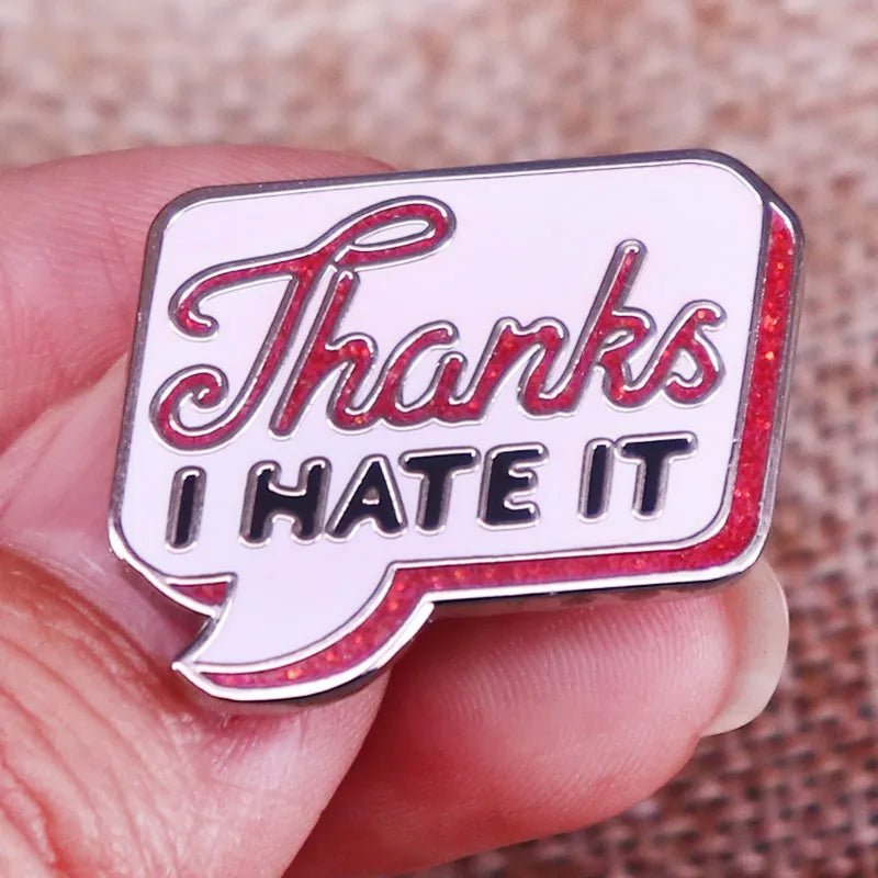 A hand holds a My Cutesy Era enamel pin shaped like a retro speech bubble reading “Thanks” in red script and “I HATE IT” in bold black on a pink background with a red border. Ships worldwide.