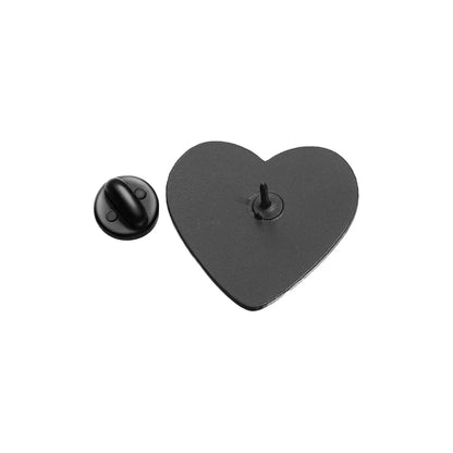 The Kiss Me Later black and white heart-shaped enamel pin, featuring a pointed post at its center, is shown beside its matching round black pin backing on a white background.
