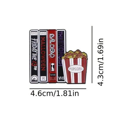 The Kiss Me Later enamel pin features three VHS tapes labeled Friday the 13th, Halloween, Evil Dead, and The Exorcist next to a popcorn bucket. Ideal for movie buffs and collectors. Size: 4.6 x 4.3 cm.