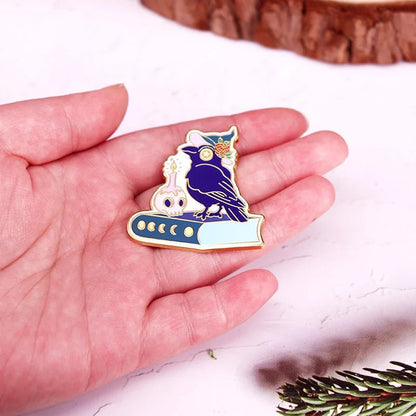 A hand holds an enamel pin showing a dark blue owl with gold outlines perched on a book, next to a small white skull topped with a flower. A wooden surface, pine needles, and a candle appear in the background.