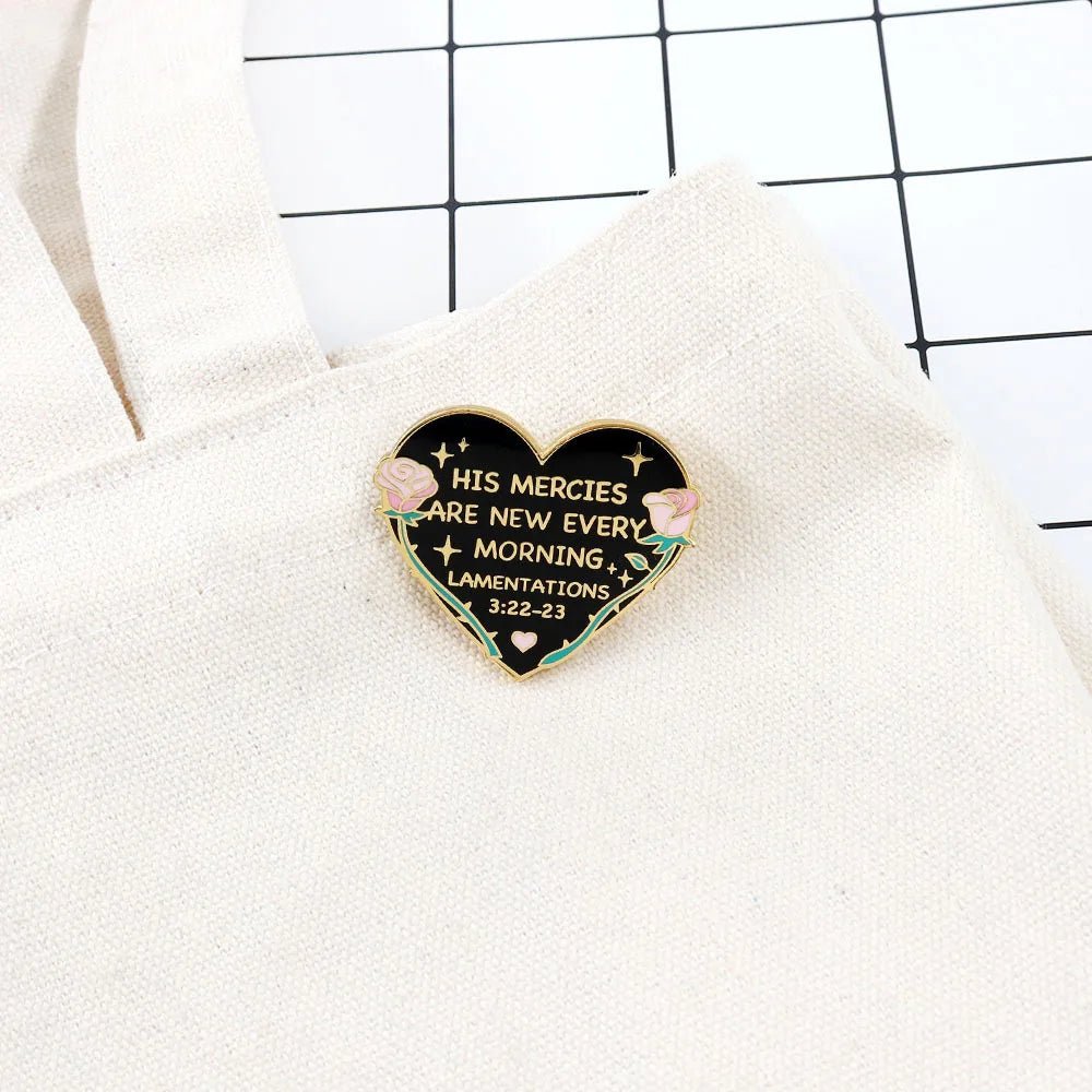 A My Cutesy Era heart-shaped enamel pin with floral accents reads “His Mercies Are New Every Morning” (Lamentations 3:22-23) and is pinned to a light fabric bag on a grid-patterned background.