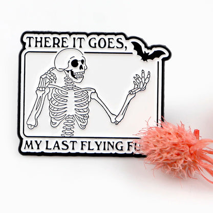 The Kiss Me Later skeleton hand enamel pin shows a smiling skeleton with one hand up, spooky flair, and THERE IT GOES, MY LAST FLYING FU text with a bat above. A pink fuzzy object partly covers the lower right text.