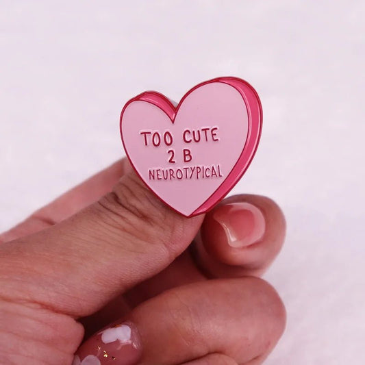 A hand with decorated nails holds a pink candy heart pin that reads TOO CUTE 2 B NEUROTYPICAL—a playful way to express neurodiversity pride.