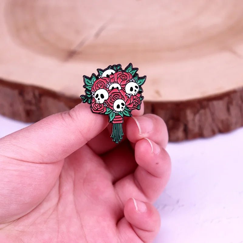 A hand holds an enamel pin shaped like a gothic bouquet of red roses with tiny white skulls among the flowers, set against a wooden background.