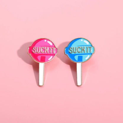 Two enamel pins on a pink background—one pink, one blue—each boldly saying SUCK IT. The perfect playful, candy-inspired accessory for those who love fun and bold style.