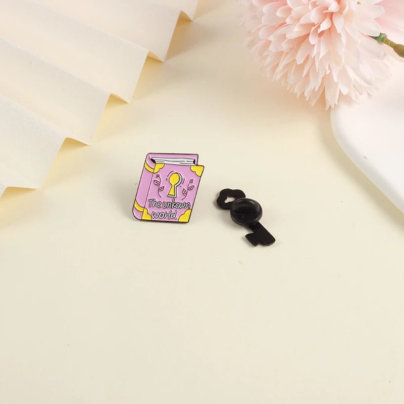 A small pink enamel book badge titled The Unknown World with a keyhole cover design, sits beside its black fastener on a cream surface near a pink flower—ideal for those who love to explore the world.