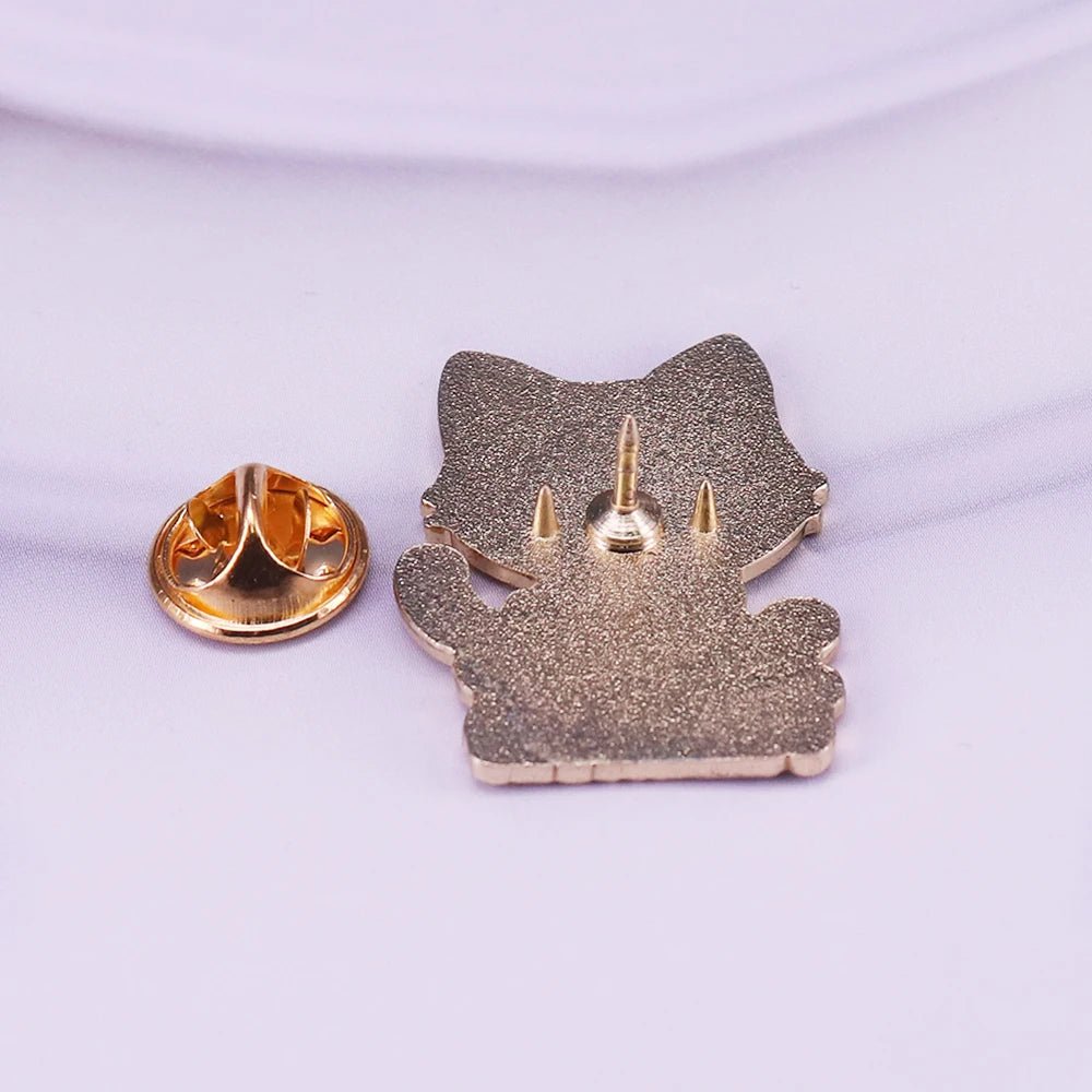 Gold cat enamel pin with three prongs is displayed next to its separate gold butterfly clutch back on a soft, light purple surface.