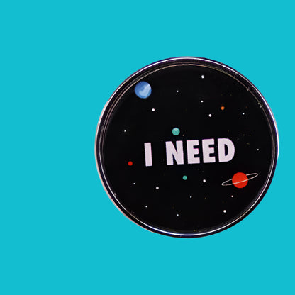 A gloved hand holds a round black metal pin from Kiss Me Later featuring small planets, stars, and I NEED in white—an on-trend accessory ideal for brooch and pin collectors.