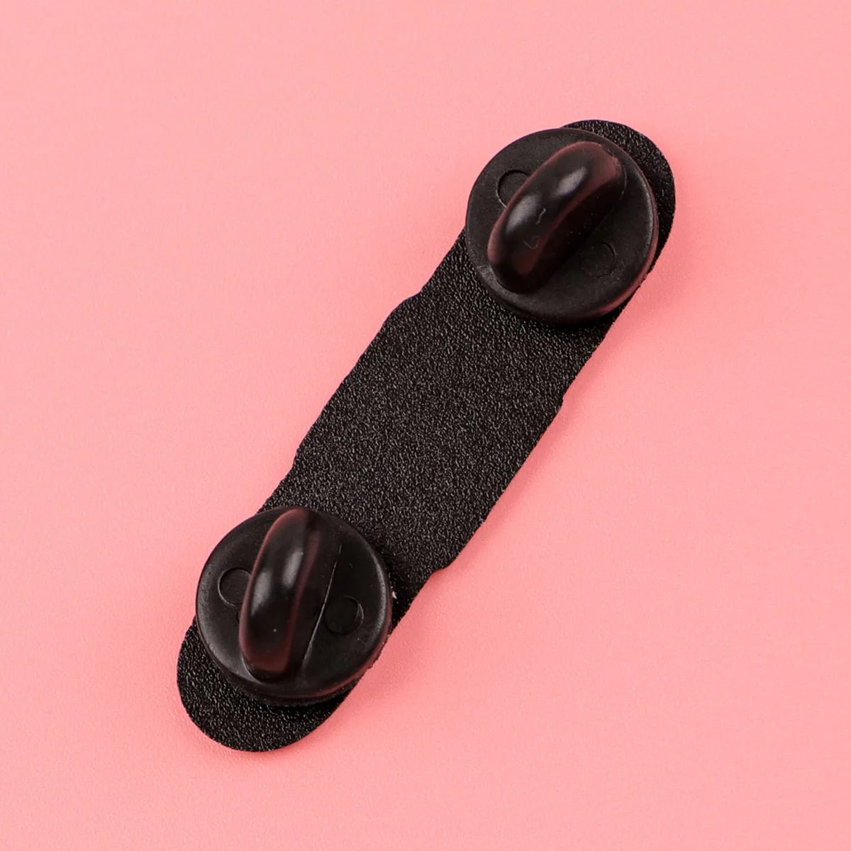 black double clasp fastener—made of fabric and plastic—sits on a pink background, perfect for holding an enamel pin or pink bandaid.