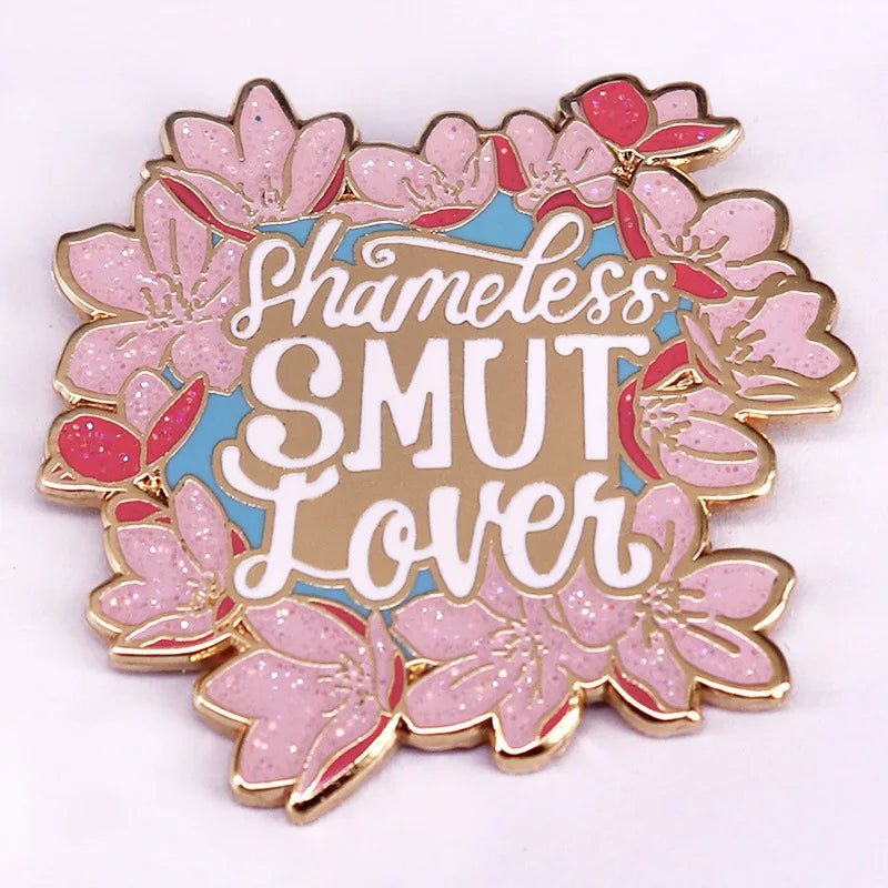 Add playful flair to your look with an enamel pin: pink flowers frame Shameless Smut Lover in bold white script on a light blue background—the perfect accessory for book lovers with a sense of fun.