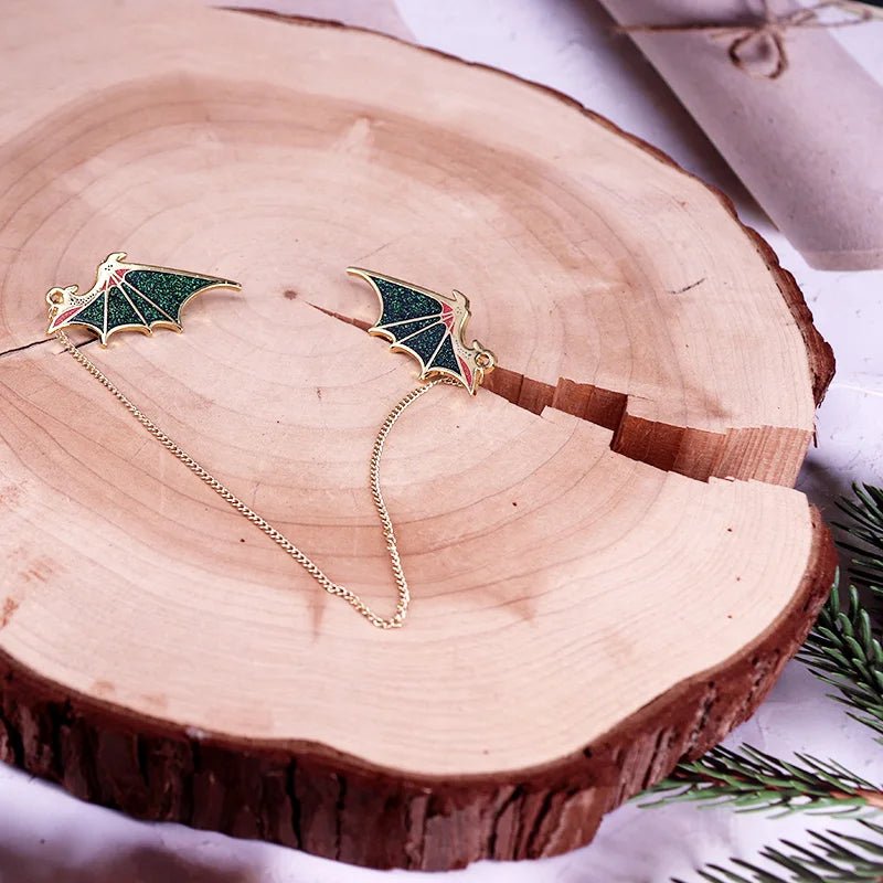 A pair of glittering green and gold wing-shaped earrings, linked by a chain, rest on a wooden slice with evergreen sprigs and a wrapped gift nearby for a festive touch.