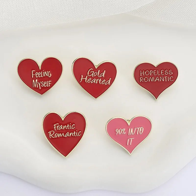 Five heart-shaped enamel pins — four red, one pink—feature phrases: Feeling Myself, Gold Hearted, Hopeless Romantic, Frantic Romantic, and 90% Into It. Displayed on a white fabric background.