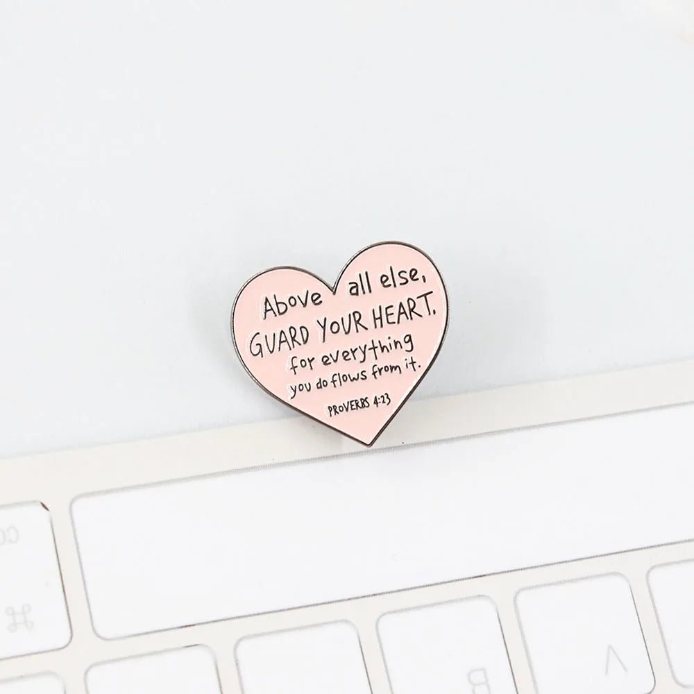 A Kiss Me Later pink love heart enamel pin reading Above all else, guard your heart, for everything you do flows from it. Proverbs 4:23 rests gracefully on a white computer keyboard.
