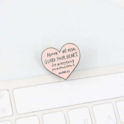 A Kiss Me Later pink love heart enamel pin reading Above all else, guard your heart, for everything you do flows from it. Proverbs 4:23 rests gracefully on a white computer keyboard.