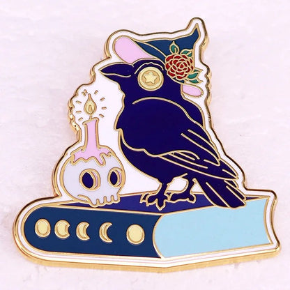 Enamel pin features a black raven in a pink witch hat with a rose, perched on a blue book, next to a skull-shaped potion bottle topped with a candle—perfect for adding whimsical charm to your collection.