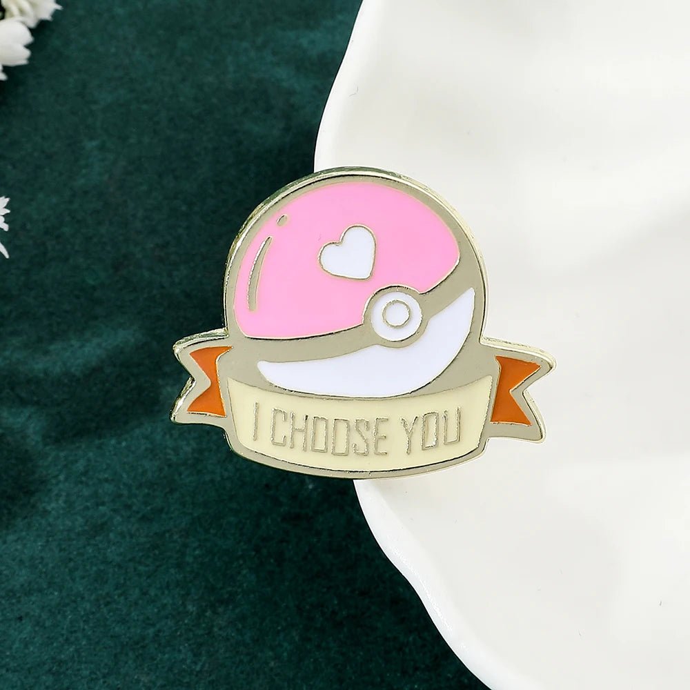 Pink Pokémon ball enamel pin features a heart on top and a banner reading I CHOOSE YOU. Shown on the edge of a white dish atop a green surface, it’s the perfect gift for any Pokémon fan!.