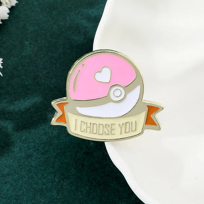 Pink Pokémon ball enamel pin features a heart on top and a banner reading I CHOOSE YOU. Shown on the edge of a white dish atop a green surface, it’s the perfect gift for any Pokémon fan!.