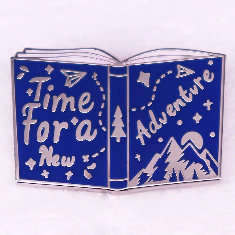 Enamel pin is shaped like an open book with blue pages, perfect for Romance Readers. It features “Time For a New Adventure,” paper planes, stars, and cute mountain and tree illustrations.