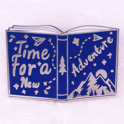 Enamel pin is shaped like an open book with blue pages, perfect for Romance Readers. It features “Time For a New Adventure,” paper planes, stars, and cute mountain and tree illustrations.