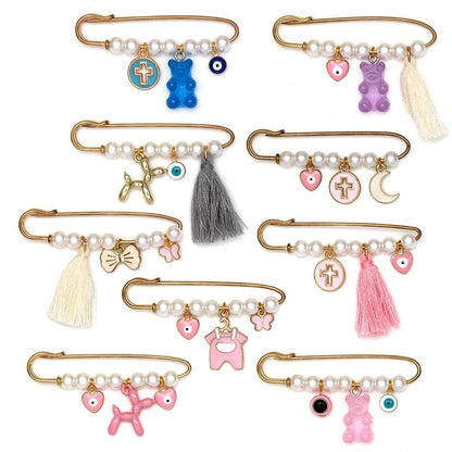Kiss Me Later presents a set of 8 decorative gold baby safety pins with pearl beads and assorted charms—evil eye, hearts, bows, and balloon dog—arranged on white. A charming brooch set, ideal as a baby shower gift.