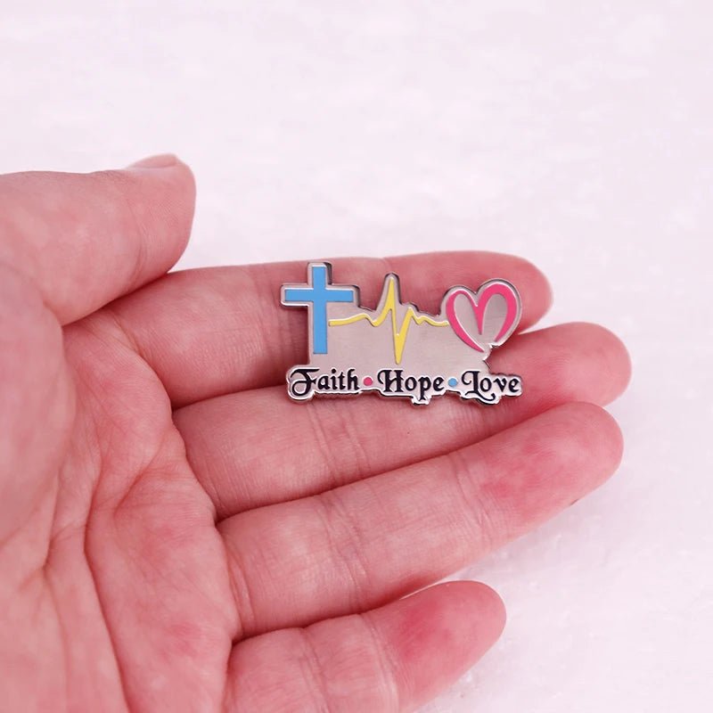 A hand holds a vibrant Kiss Me Later Faith Hope Love badge with a blue cross, yellow heartbeat line, and pink heart on a light pink background.