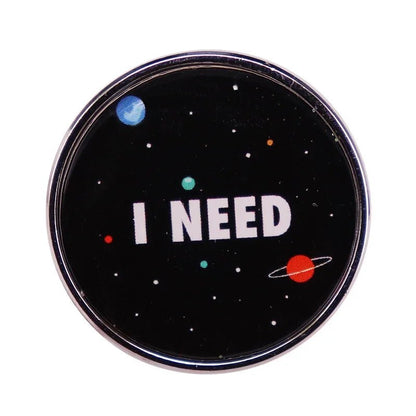 A round black metal pin from Kiss Me Later, featuring planets and stars with bold white I NEED text at the center—perfect for fans of trendy fashion accessories.