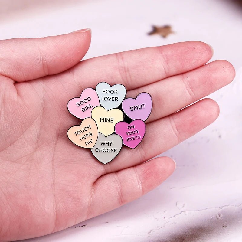 A hand displays seven colourful love hearts featuring enamel pins like Book Lover and Smut, with phrases such as GOOD GIRL and ON YOUR KNEES—ideal bookish flair for pin collectors.