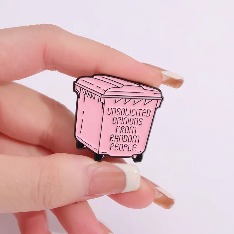 A hand holds a pink enamel pin shaped like a trash bin, featuring the words UNSOLICITED OPINIONS FROM RANDOM PEOPLE on its side.