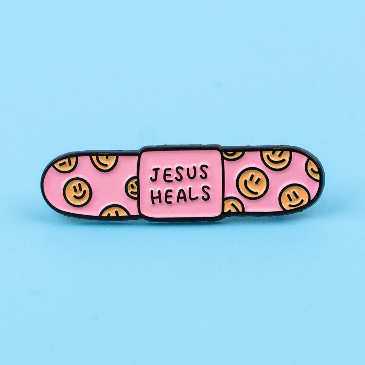 pink bandaid enamel pin with yellow smiley faces on the ends and JESUS HEALS in the center, displayed on a light blue background.