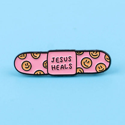 pink bandaid enamel pin with yellow smiley faces on the ends and JESUS HEALS in the center, displayed on a light blue background.