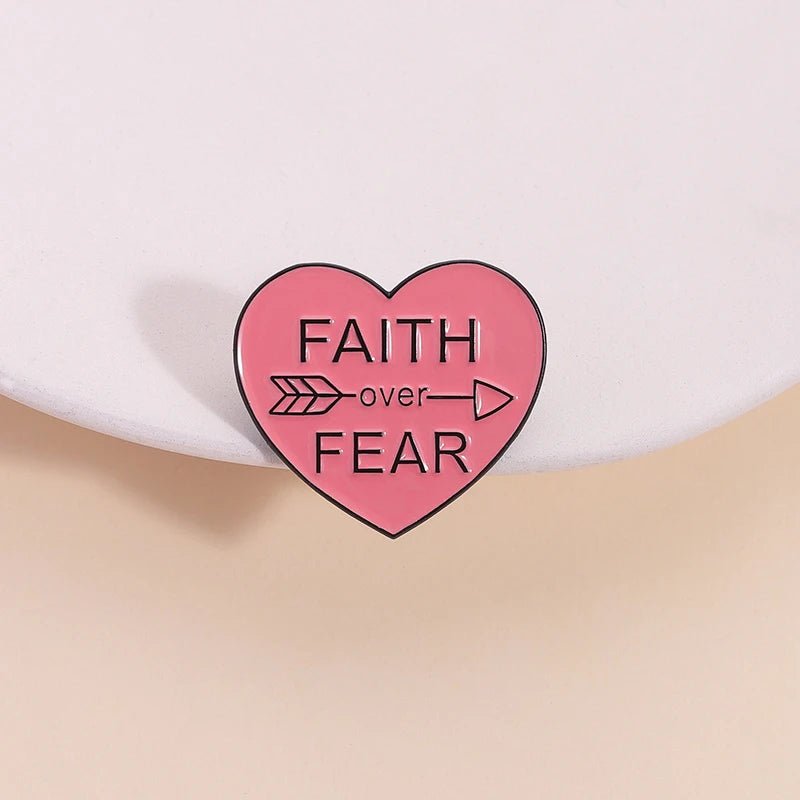 Pink enamel pin, shaped like a love heart with FAITH OVER FEAR and a right-pointing arrow, is shown on a light-colored surface.