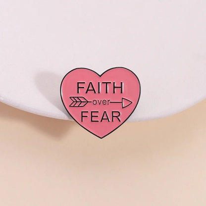 Pink enamel pin, shaped like a love heart with FAITH OVER FEAR and a right-pointing arrow, is shown on a light-colored surface.