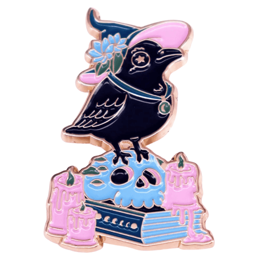 Gothic Book Crow enamel pin features a black bird in a purple witch hat with a blue flower, perched on a blue skull and book, surrounded by pink melting candles, all outlined in gold.