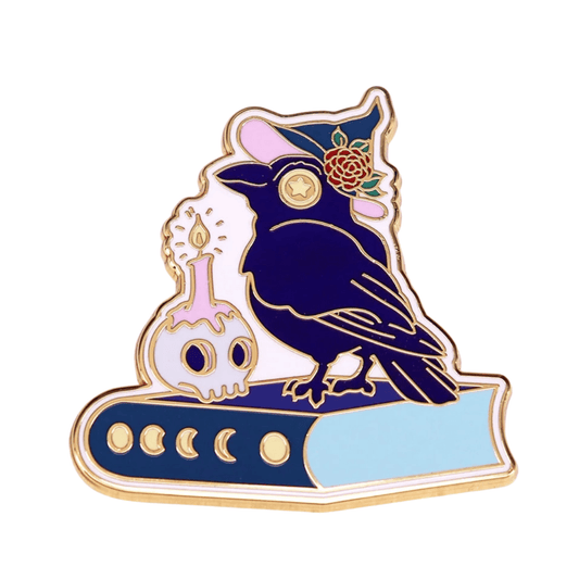 Enamel pin features a black raven in a witch hat with flowers, perched on a blue book next to a skull candle. Gold outlines and colorful accents give this pin a magical, mysterious look.
