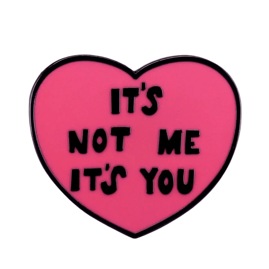A heart-shaped pink enamel pin  featuring bold black letters that read ITS NOT ME ITS YOU, stands out against a plain white background.