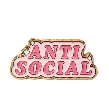 A gold-trimmed enamel pin by Kiss Me Later featuring ANTI SOCIAL in bold pink letters on a light pink background—a unique mental health accessory for self-expression.