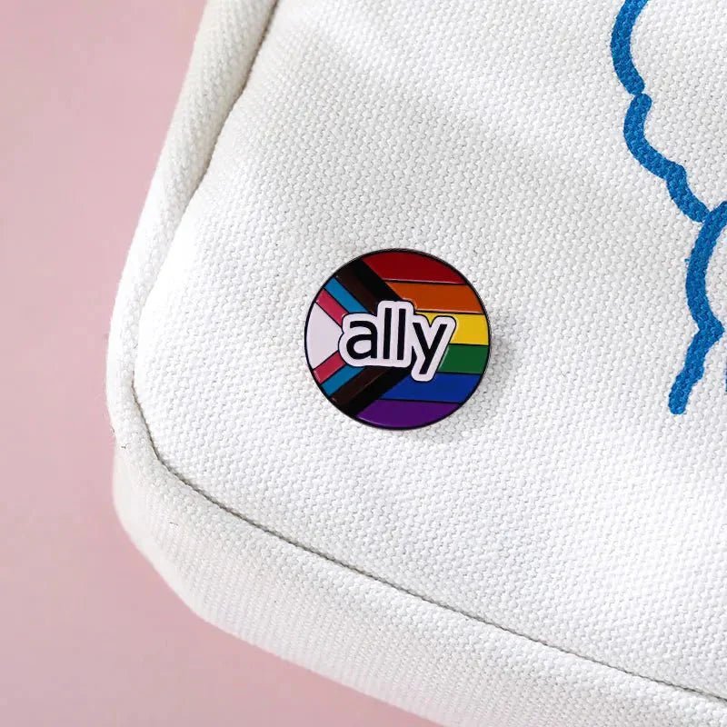 PRIDE FLAG round ally enamel pin, featuring ally over a rainbow and progress pride flag, is attached to a cream fabric bag with blue trim on a pink backdrop. Enjoy worldwide shipping on this meaningful accessory.