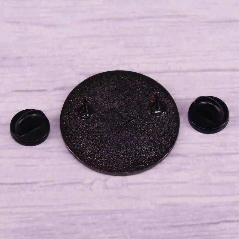 A round, textured black metal pin with two prongs is displayed on a light wood surface, alongside two removable black rubber pin backs—ideal for your collection. Worldwide shipping available.
