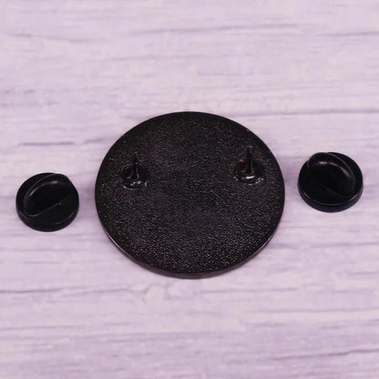 A round, textured black metal pin with two prongs is displayed on a light wood surface, alongside two removable black rubber pin backs—ideal for your collection. Worldwide shipping available.