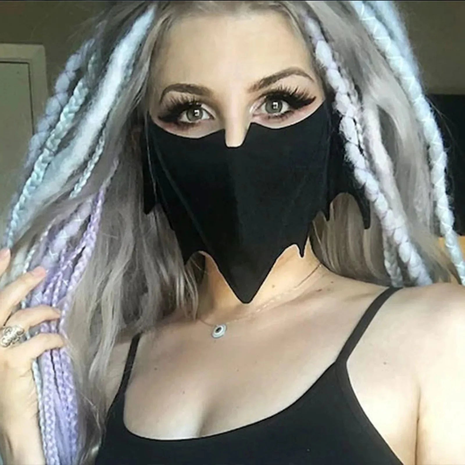 A woman with long, light purple and gray braids looks into the camera, wearing bold eye makeup, a dramatic black bat mask by Gay Skate, and a black tank top. Her reusable cloth mask adds a unique, mysterious touch to her striking look.