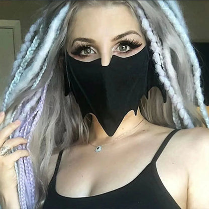 A woman with long, light purple and gray braids looks into the camera, wearing bold eye makeup, a dramatic black bat mask by Gay Skate, and a black tank top. Her reusable cloth mask adds a unique, mysterious touch to her striking look.